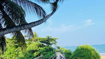 A concrete pathway with metal railings leading to a beach, surrounded by lush greenery and palm trees.