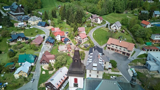 Eine Vogelperspektive eines Dorfes mit verschiedenen Gebäuden, darunter eine Kirche mit einem markanten Kirchturm, umgeben von Wohnhäusern und einem Skigebiet.
