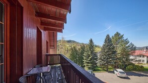 Ein Holzbalkon mit Tisch und Stühlen, mit Blick auf einen Parkplatz und eine grüne Landschaft mit Bäumen und Gebäuden.