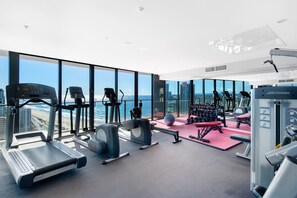A modern gym with various exercise equipment, large windows offering ocean views, and a high ceiling. - Stunning High Level Deluxe Apartment at Rhapsody (Surfers Paradise)