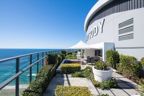 A modern building with a clear view of the ocean, a balcony with outdoor seating, and a large potted plant. - Stunning High Level Deluxe Apartment at Rhapsody (Surfers Paradise)