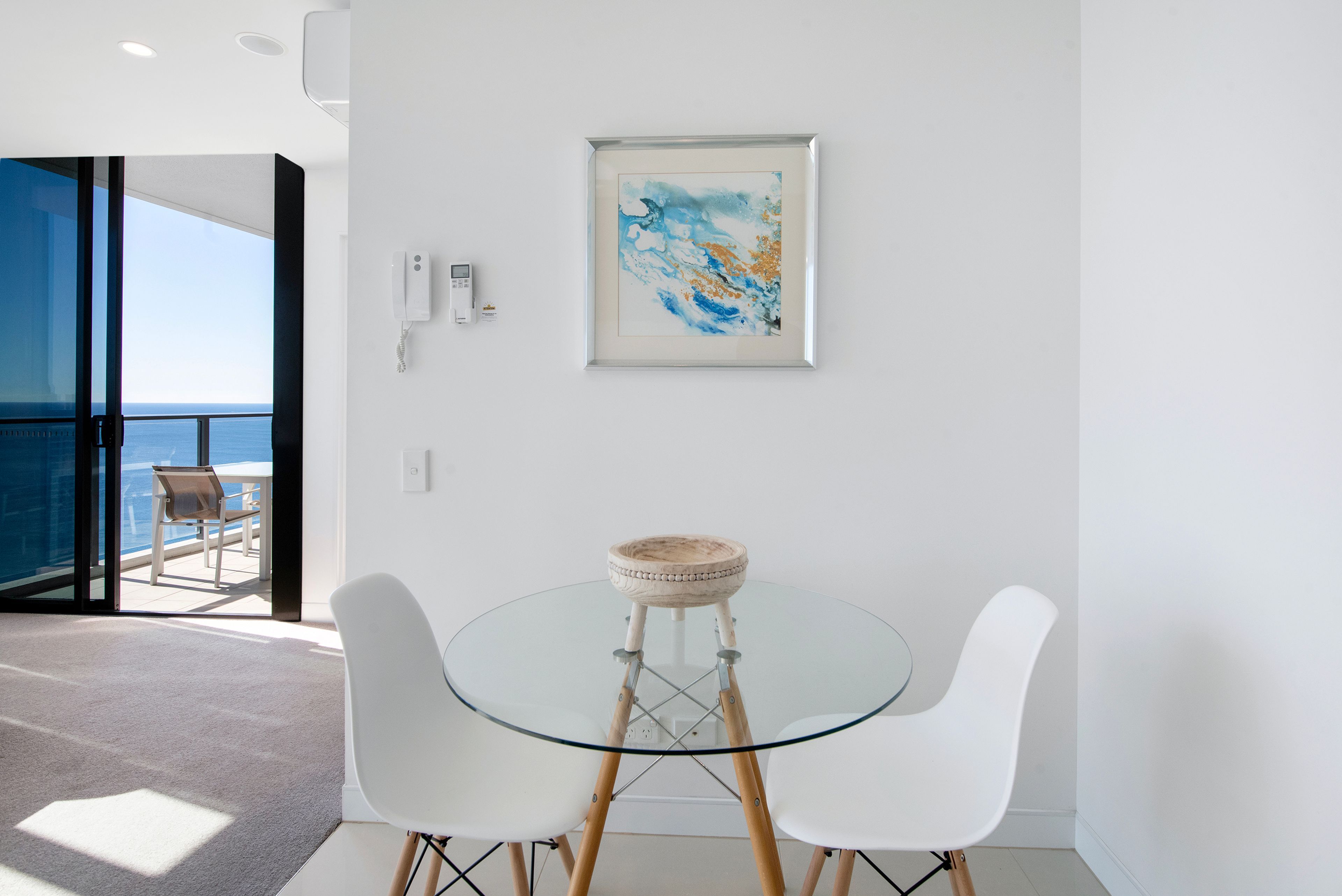 Quarto moderno com mesa de vidro, duas cadeiras brancas, um quadro abstrato emoldurado e vista para o mar através de uma porta aberta.