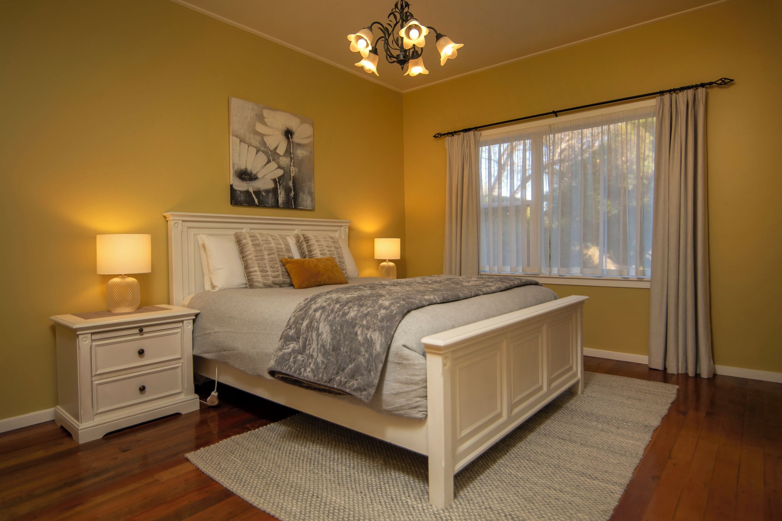 A bedroom with a bed, nightstands, a chandelier, a window with curtains, and a painting on the wall.