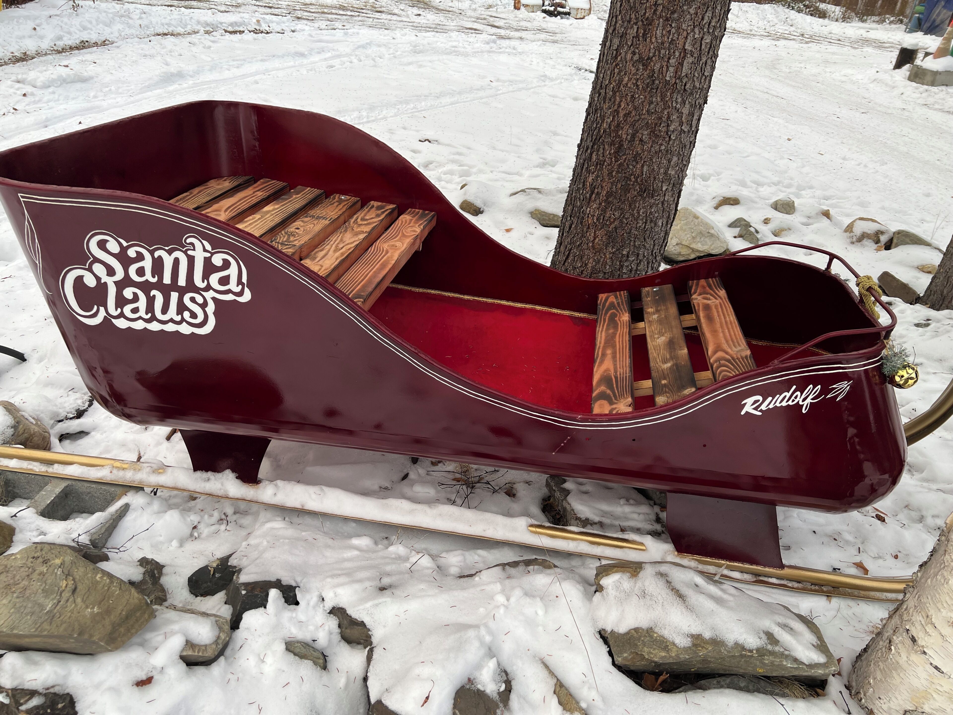 A red sleigh with 'Santa Claus' and 'Rudolf' written on it, placed on a snowy surface with rocks and a tree nearby.
