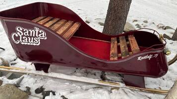 A red sleigh with 'Santa Claus' and 'Rudolf' written on it, placed on a snowy surface with rocks and a tree nearby.