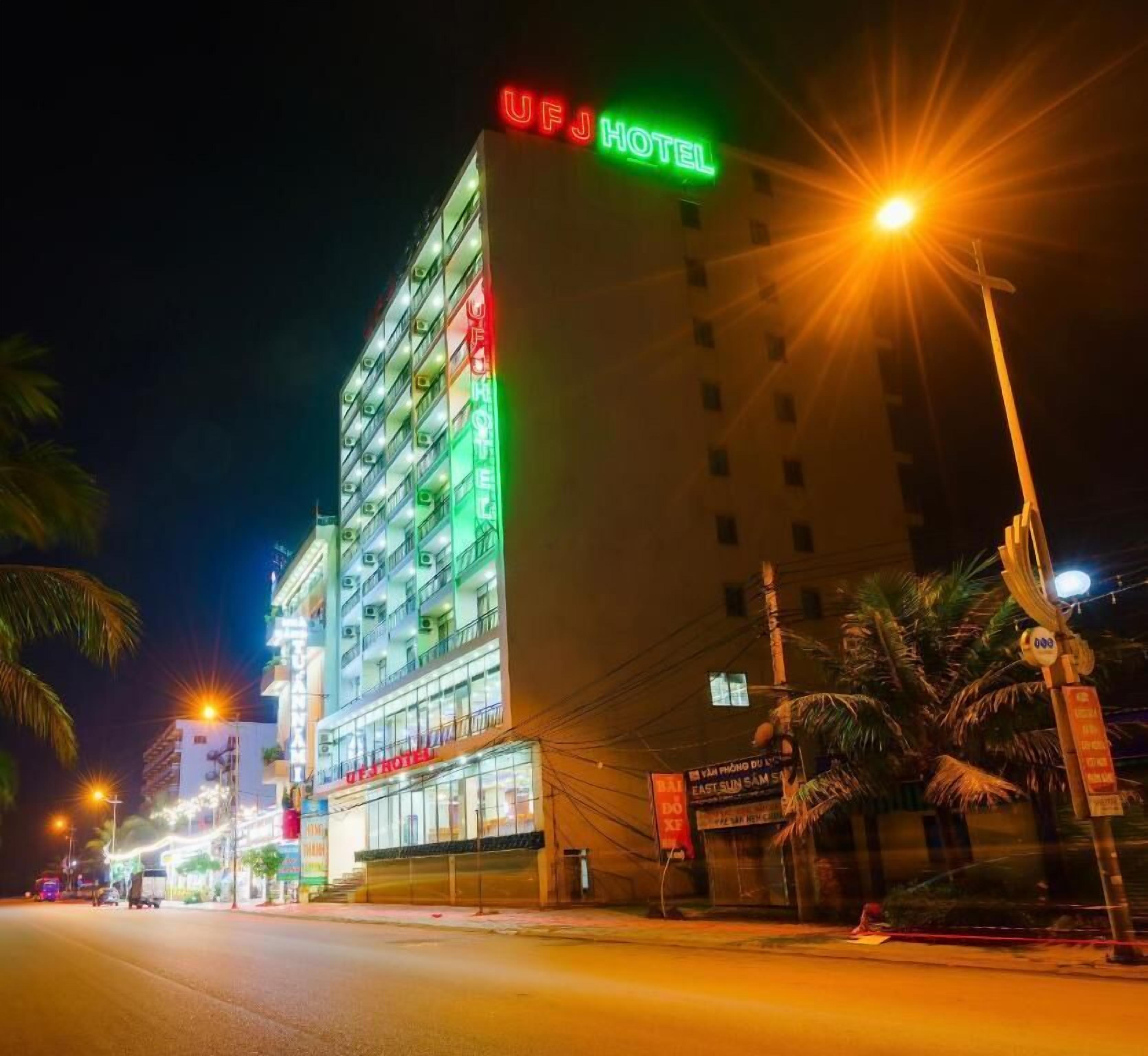 Un hotel de varios pisos con señalización iluminada, una entrada visible y una vista a la calle con palmeras y farolas.