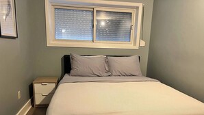 Et soveværelse med en seng, et natbord, et vindue med persienner og en vægmonteret lampe.