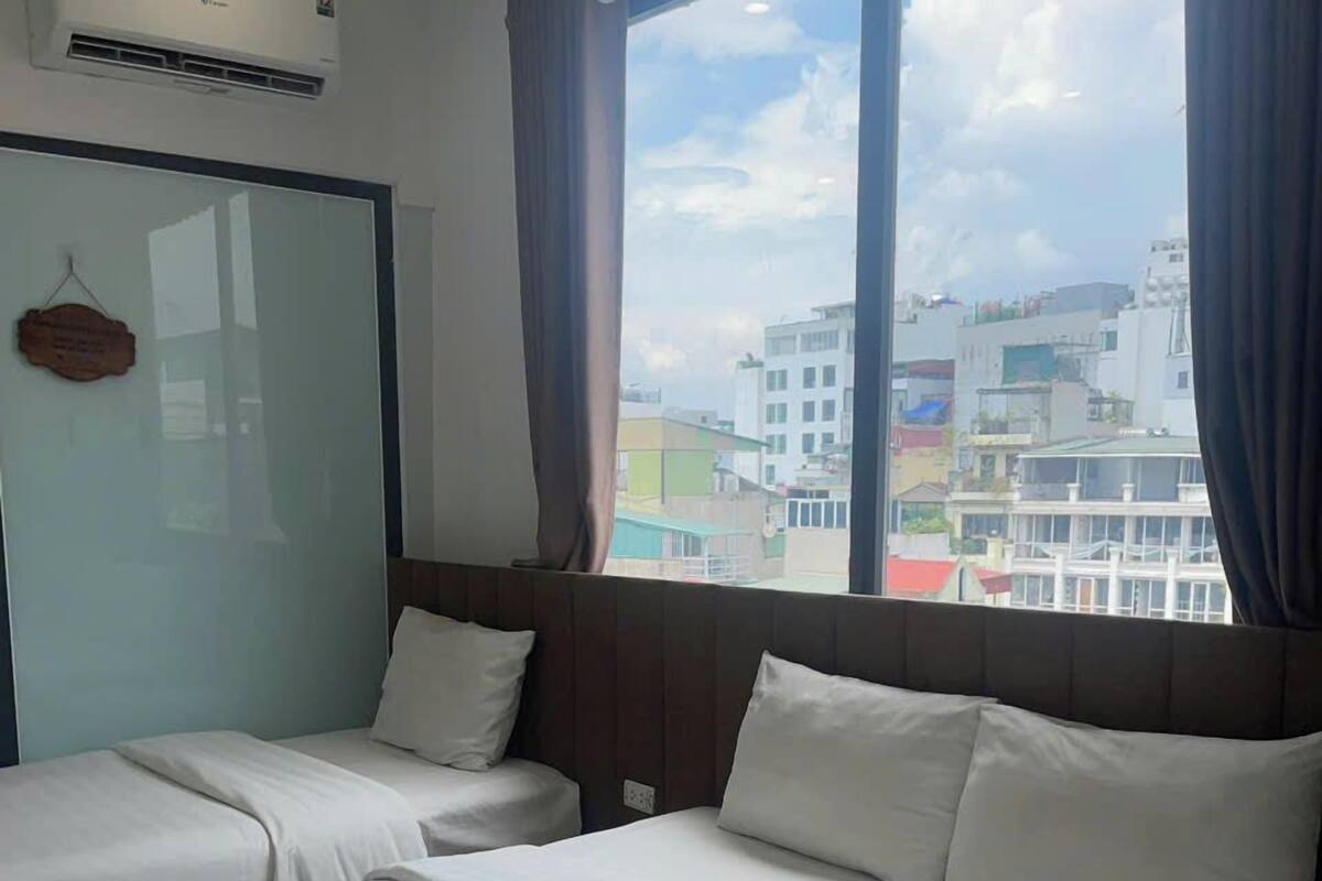 Habitación de hotel con dos camas, una ventana con cortinas, un equipo de aire acondicionado y vistas a edificios.