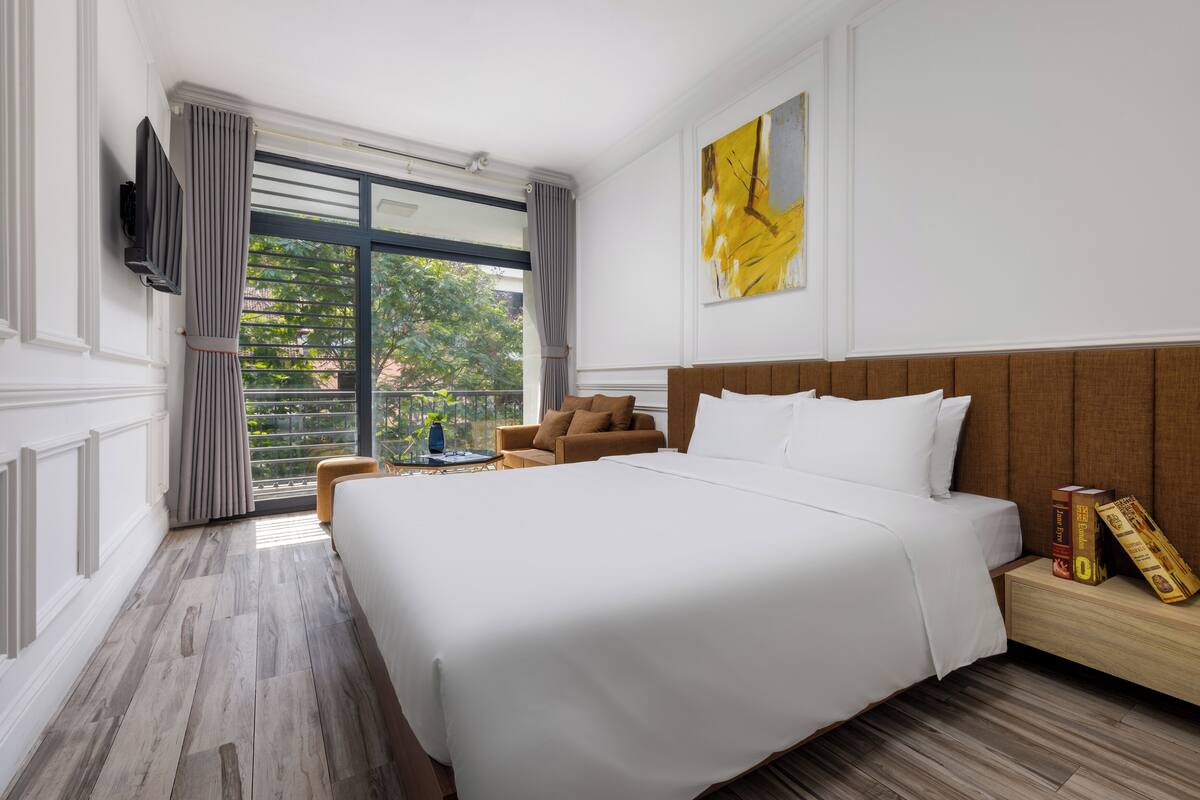 Habitación de hotel con una cama grande, cabecera de madera, televisor de pantalla plana montado en la pared, una mesita con un florero y vistas a zonas verdes por la ventana.