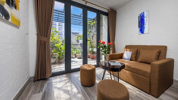 A modern living room with a brown sofa, a round ottoman, and a coffee table. There is a large window with curtains, a potted plant, and a painting on the wall. - Hanoi Old Quarter Hotel and Spa (Hanoi)