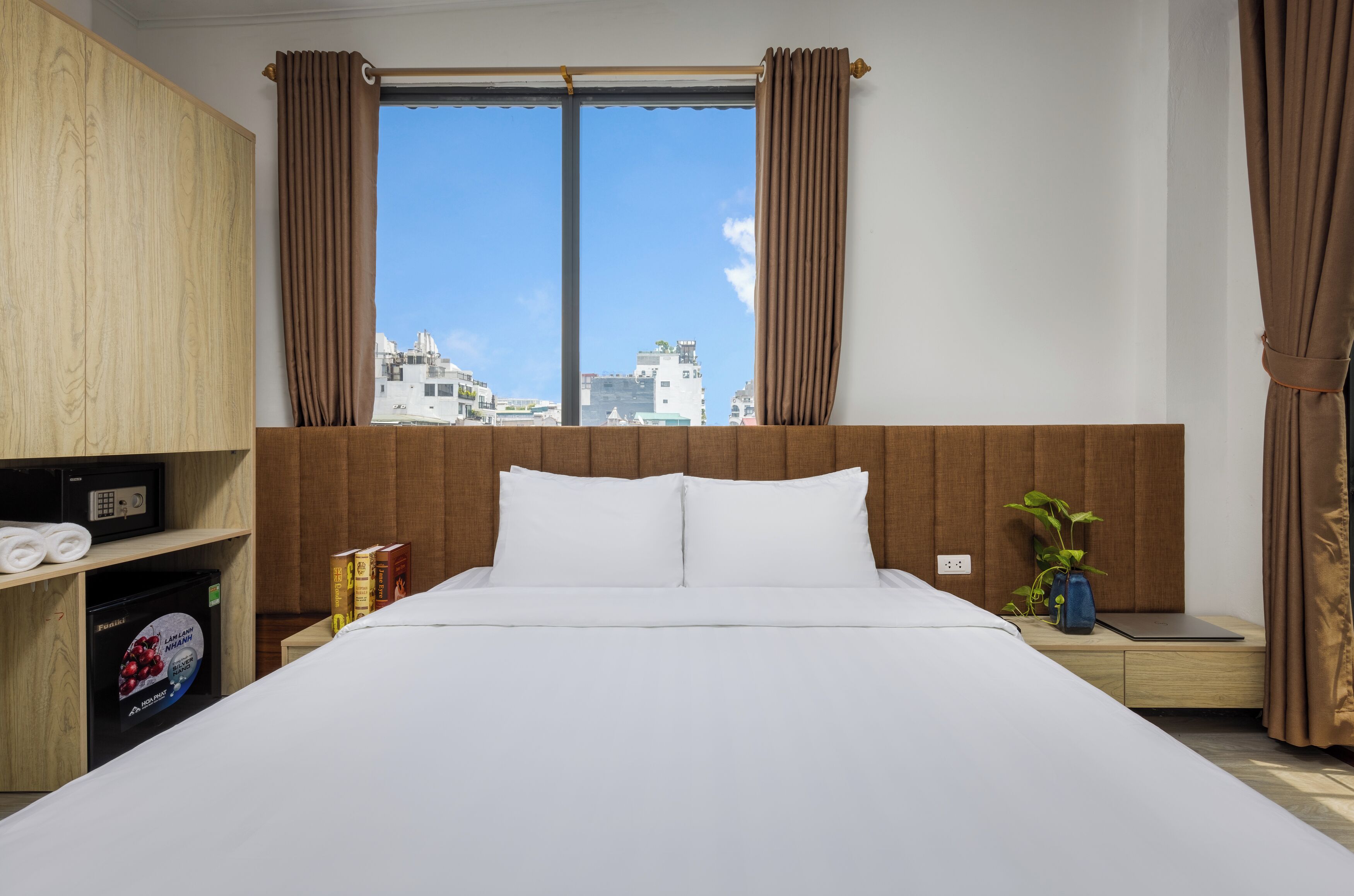 A hotel room with a large bed, wooden headboard, a view of buildings through a window, a bedside table with a plant, and a shelf with a microwave and books.