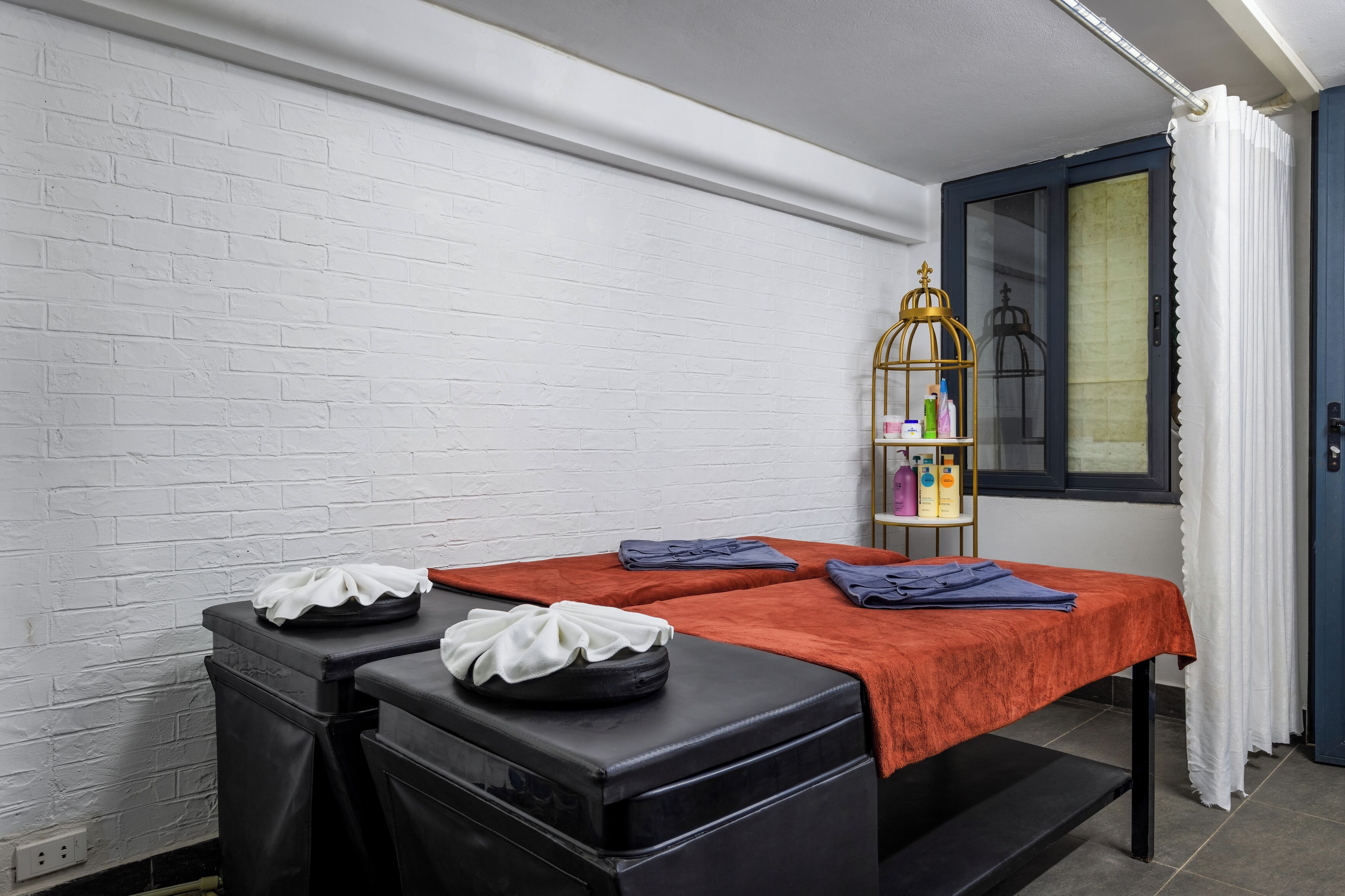 A room with two massage tables, a white brick wall, a window with a curtain, and a shelf with toiletries.
