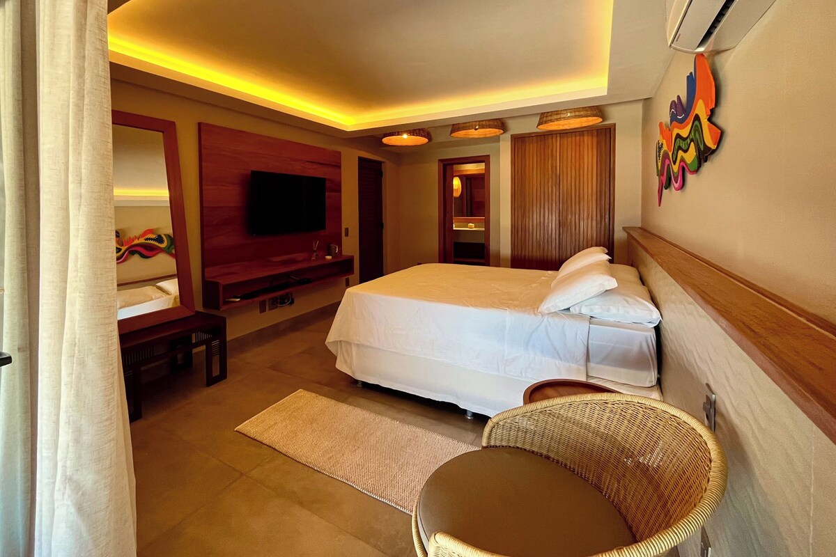 A hotel room with a bed, a television, a mirror, a wall-mounted air conditioner, and a wicker chair.
