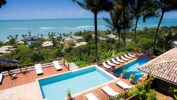A pool with lounge chairs, surrounded by tropical vegetation and overlooking a coastal area with houses. - Baixu Village (Porto Seguro)