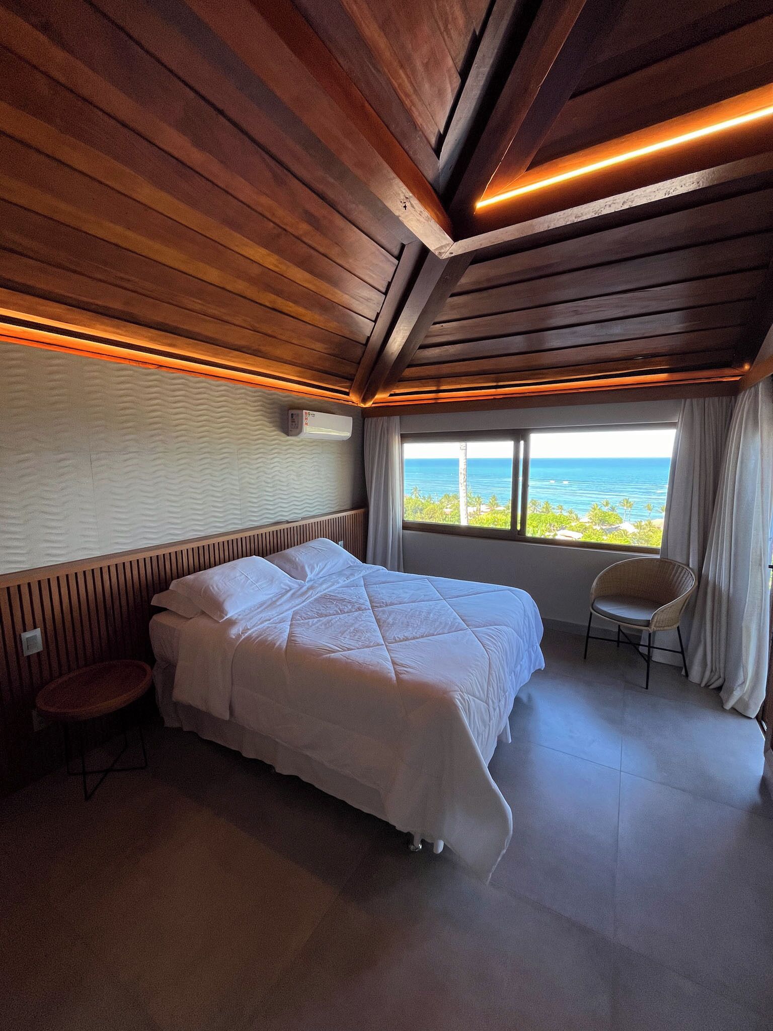 A hotel room with a bed, a chair, a window with a view of the ocean, and wooden ceiling beams.