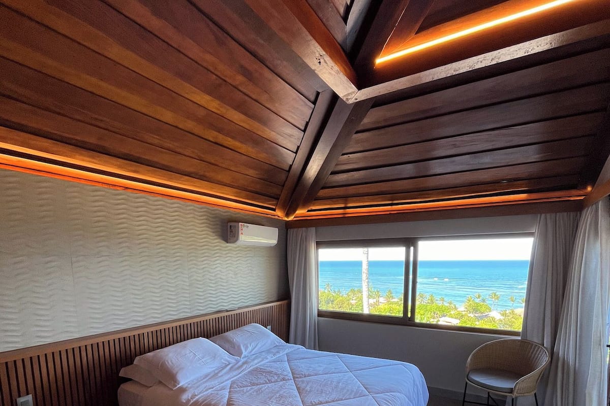 A hotel room with a bed, a chair, a window with a view of the ocean, and wooden ceiling beams.