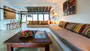 A living room with a sofa, a coffee table with magazines, a dining area with chairs, and a kitchen in the background. - Baixu Village (Porto Seguro)