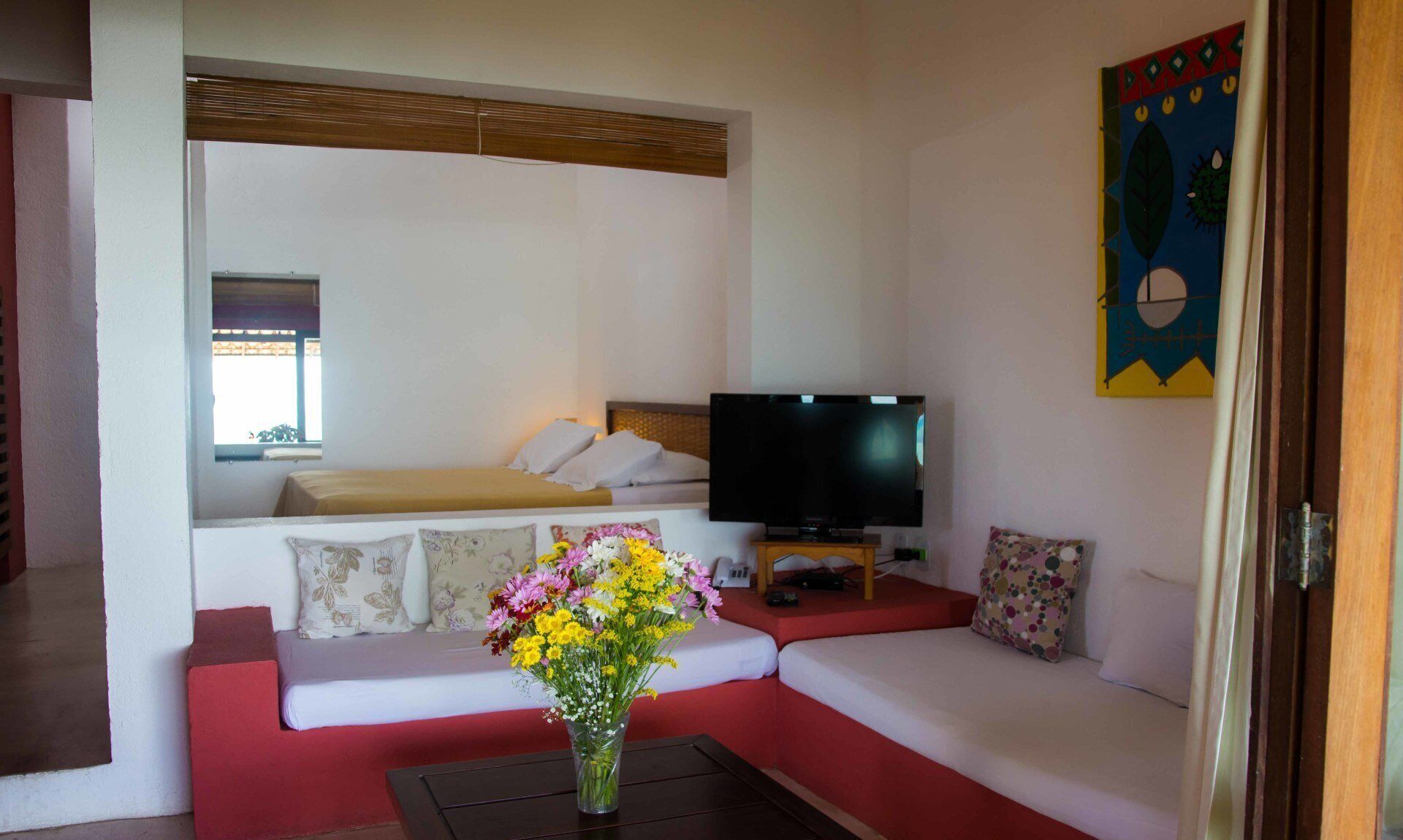 A hotel room with a bed, a TV, a sofa, a small table with a vase of flowers, and a colorful wall decoration.