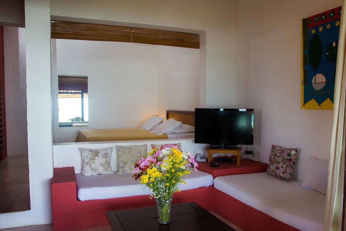 A hotel room with a bed, a TV, a sofa, a small table with a vase of flowers, and a colorful wall decoration.