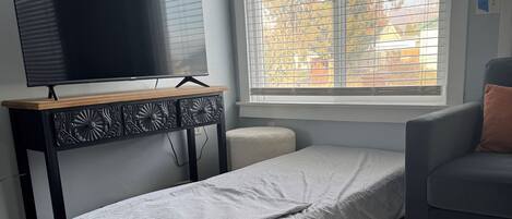 A small room with a bed, a chair, a TV on a wooden stand, and a window with blinds.