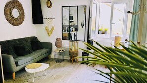 A modern living room with a grey sofa, a round mirror, a macrame wall hanging, and a large window with a view of the outdoors.