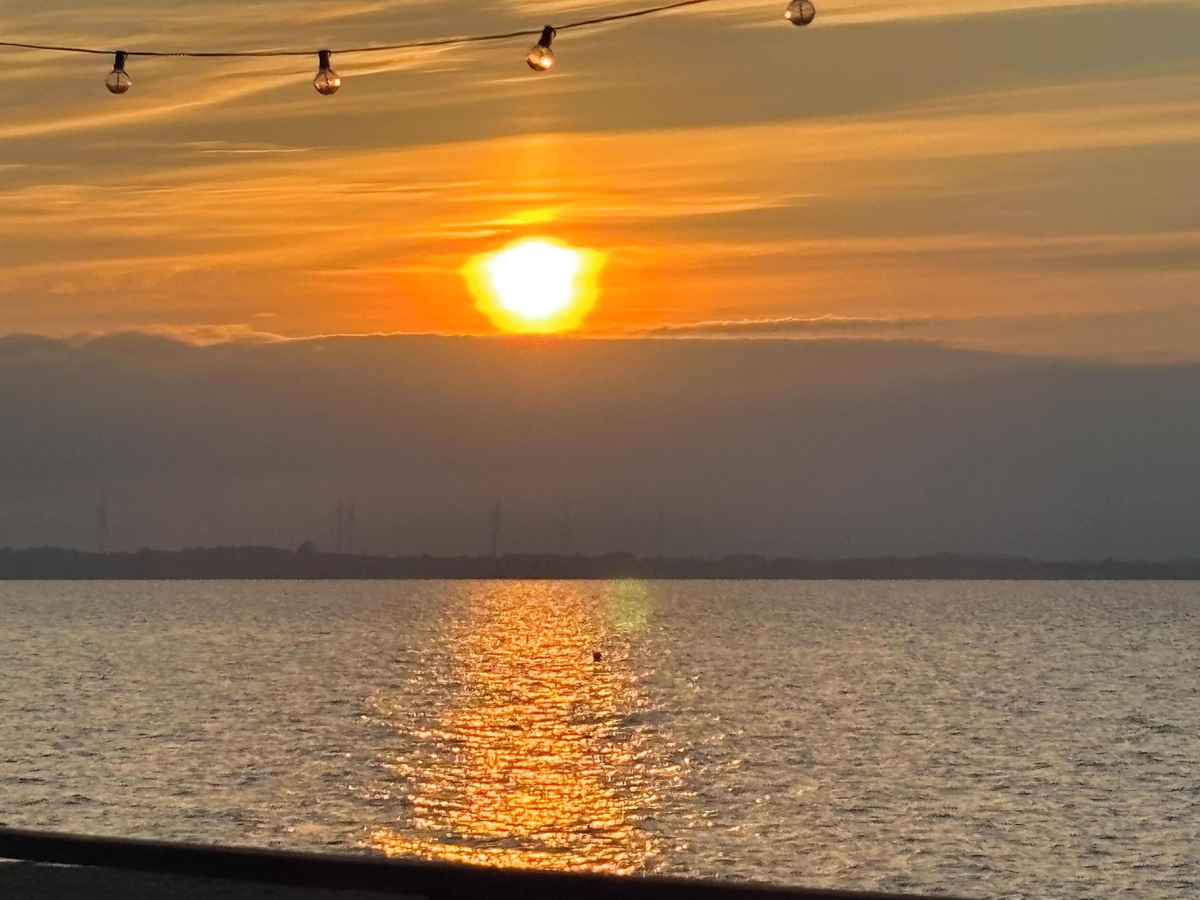 A sunset over a body of water with a distant shoreline and string lights.