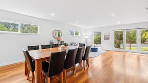 A spacious dining area with a wooden table and chairs, large windows, and a view of greenery. - Surphy G (Anglesea)
