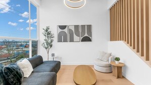 Una sala de estar moderna con un sofá, una mesa de centro y vistas a un paisaje urbano a través de amplios ventanales.