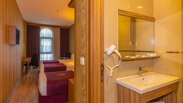 Ein Hotelzimmer mit Holzboden, einem Bett mit rotem Kopfteil, einer Sitzecke mit Sessel und Tisch, einem Badezimmer mit Waschbecken und Spiegel sowie einem Fenster mit Vorhängen.