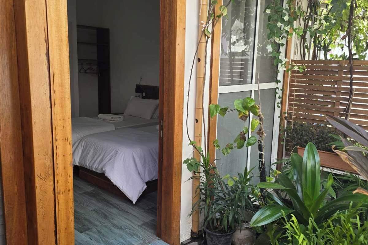 An open wooden door leading to a bedroom with a bed and a ceiling fan. There are potted plants and a wooden deck outside.