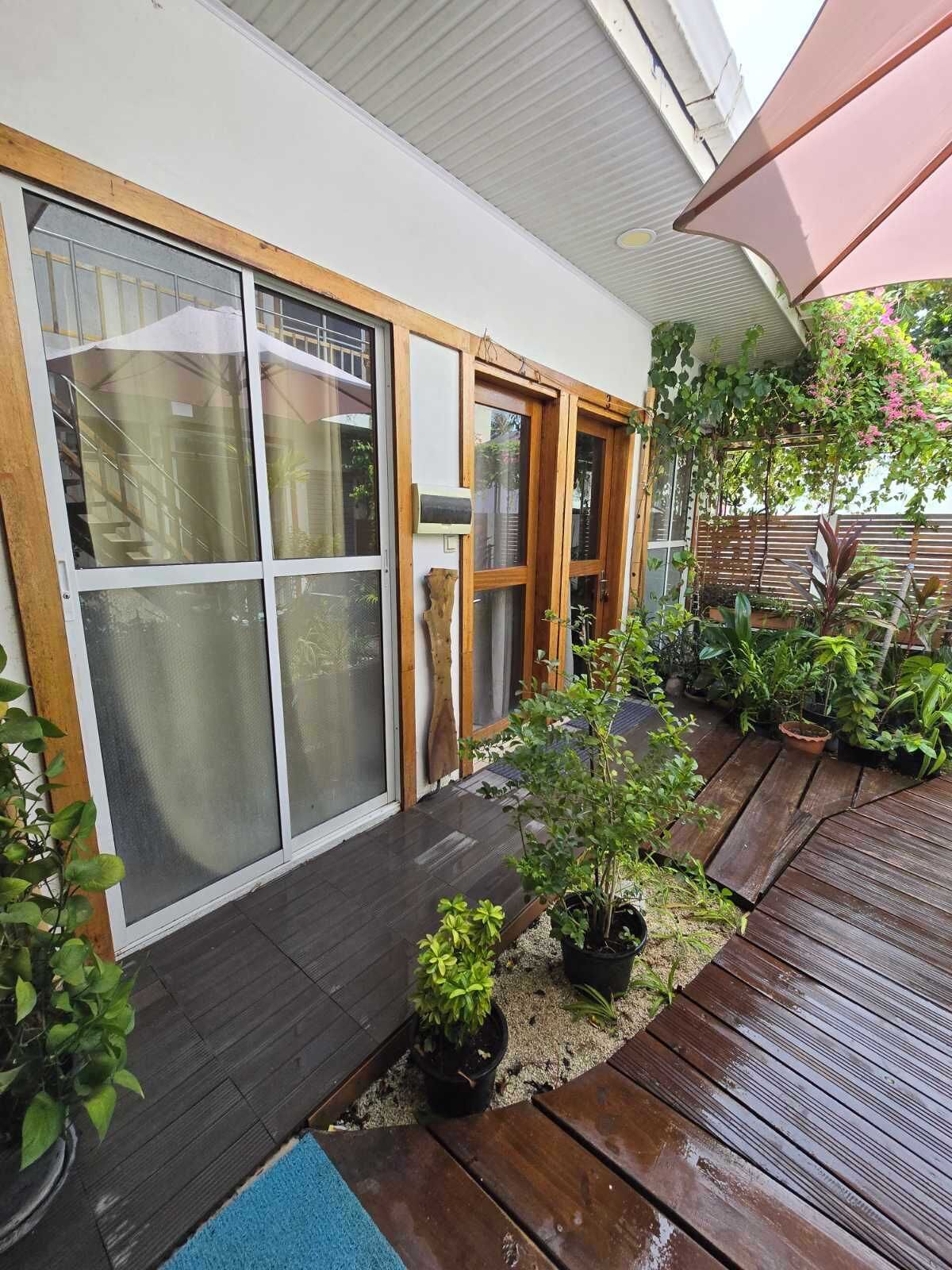 A wooden deck with potted plants, a blue mat, and a sliding glass door leading to a covered patio area.