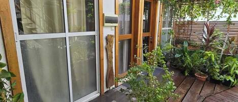 A wooden deck with potted plants, a blue mat, and a sliding glass door leading to a covered patio area.