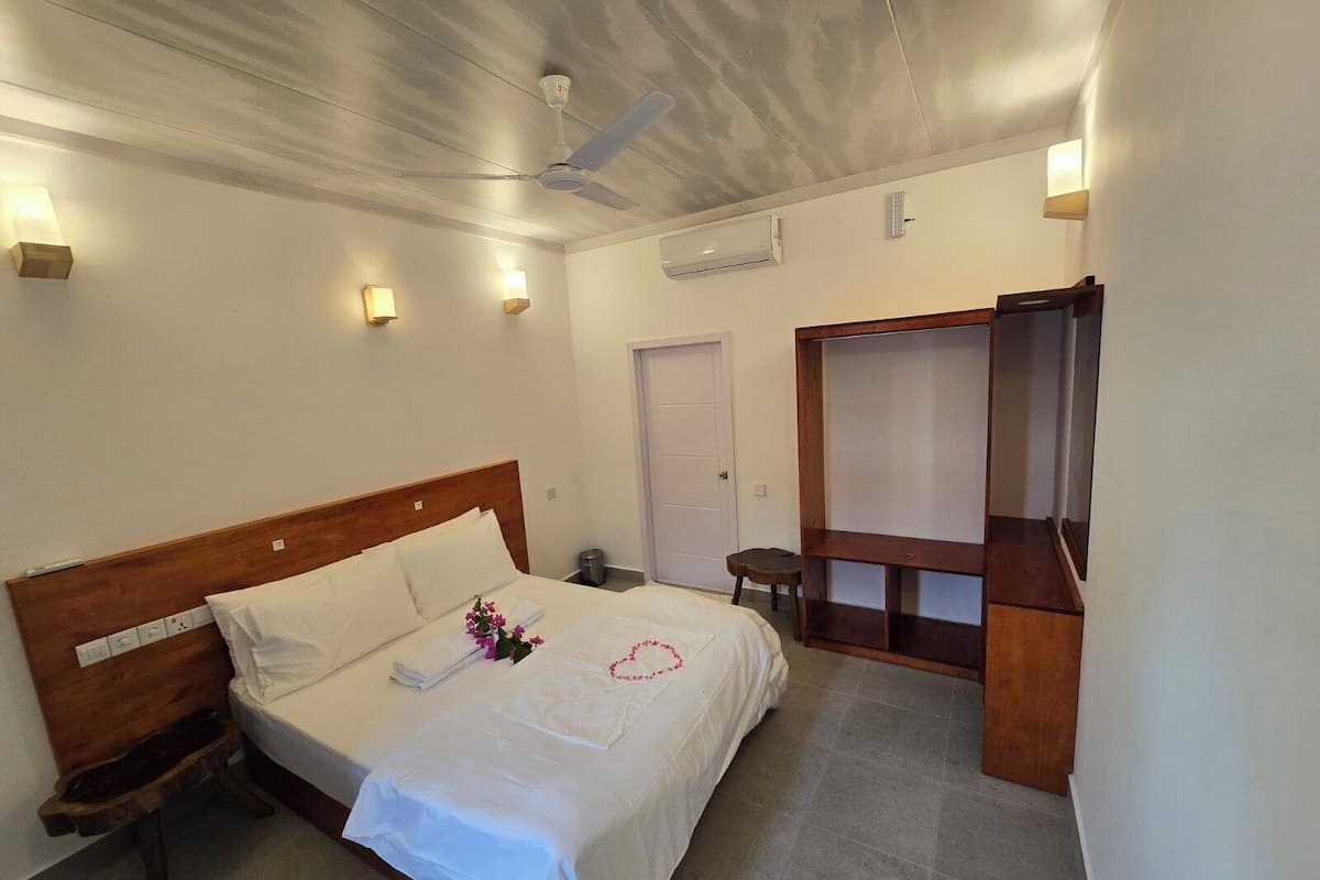 A hotel room with a bed, bedside table, nightstand, ceiling fan, wall-mounted air conditioner, and a wooden wardrobe.