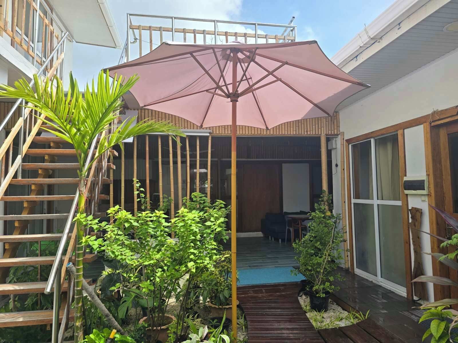 A covered outdoor area with a wooden deck, a table, and chairs, surrounded by various plants and a large umbrella.