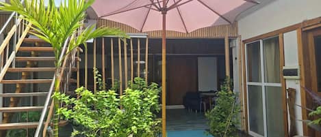 A covered outdoor area with a wooden deck, a table, and chairs, surrounded by various plants and a large umbrella.