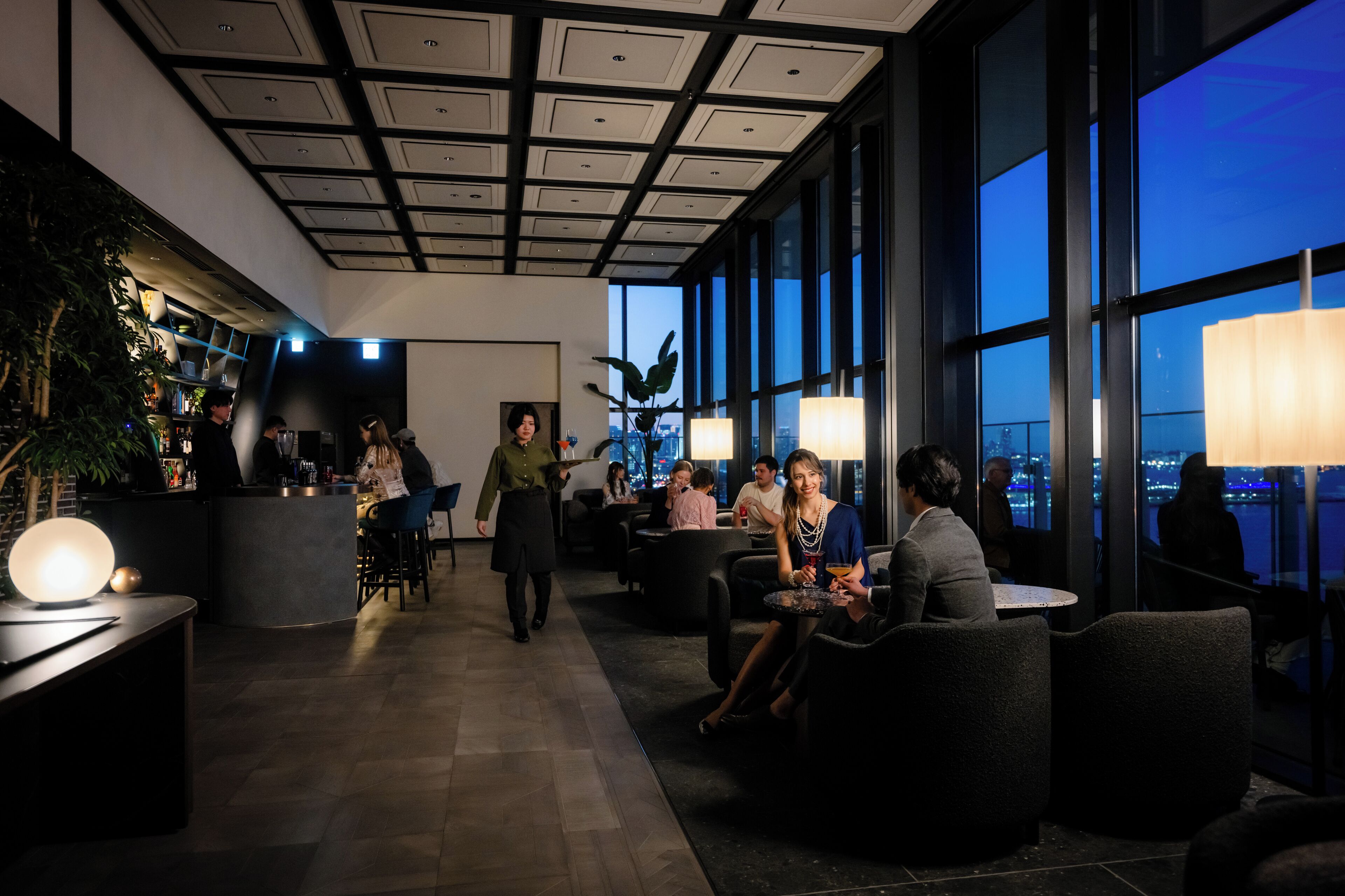 A modern lounge area with a bar, seating, and large windows offering a city view.