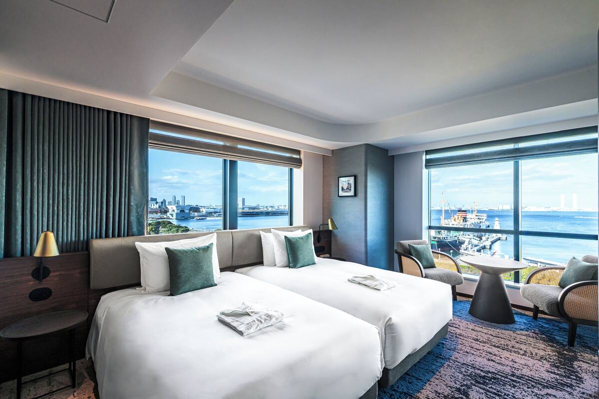 Une chambre d’hôtel avec deux lits, un coin salon avec une table et de grandes fenêtres donnant sur la ville.