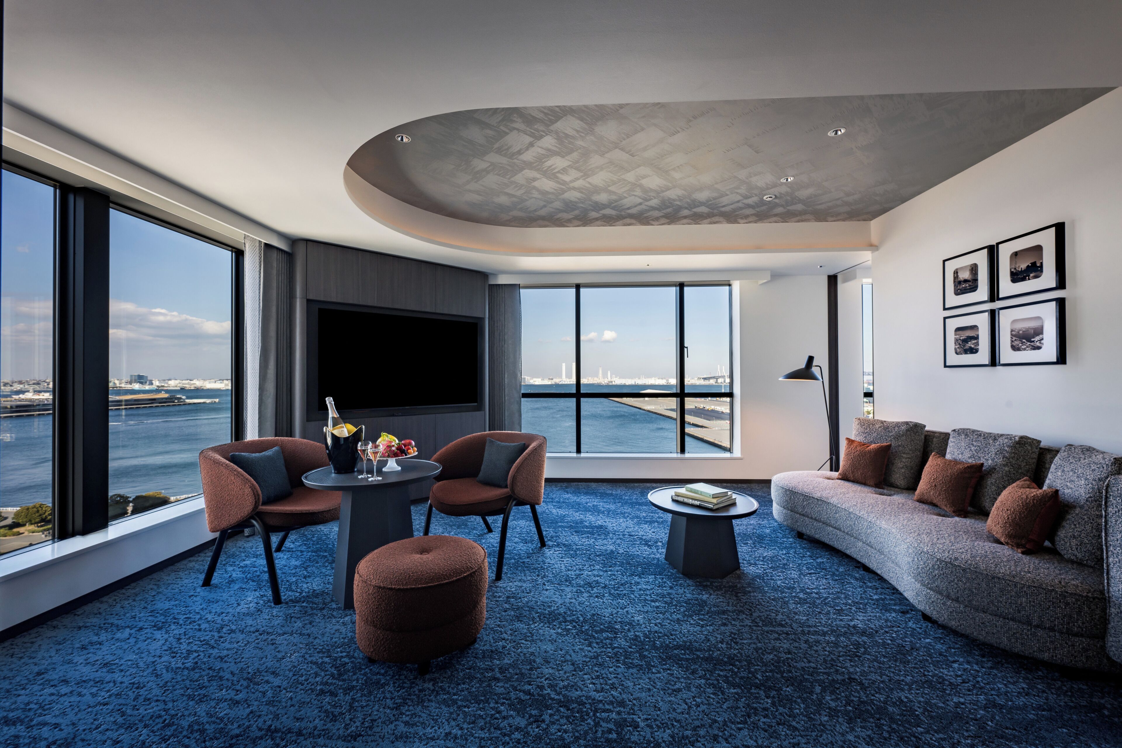 A modern living room with a large TV, comfortable seating, a coffee table, and a view of the water.
