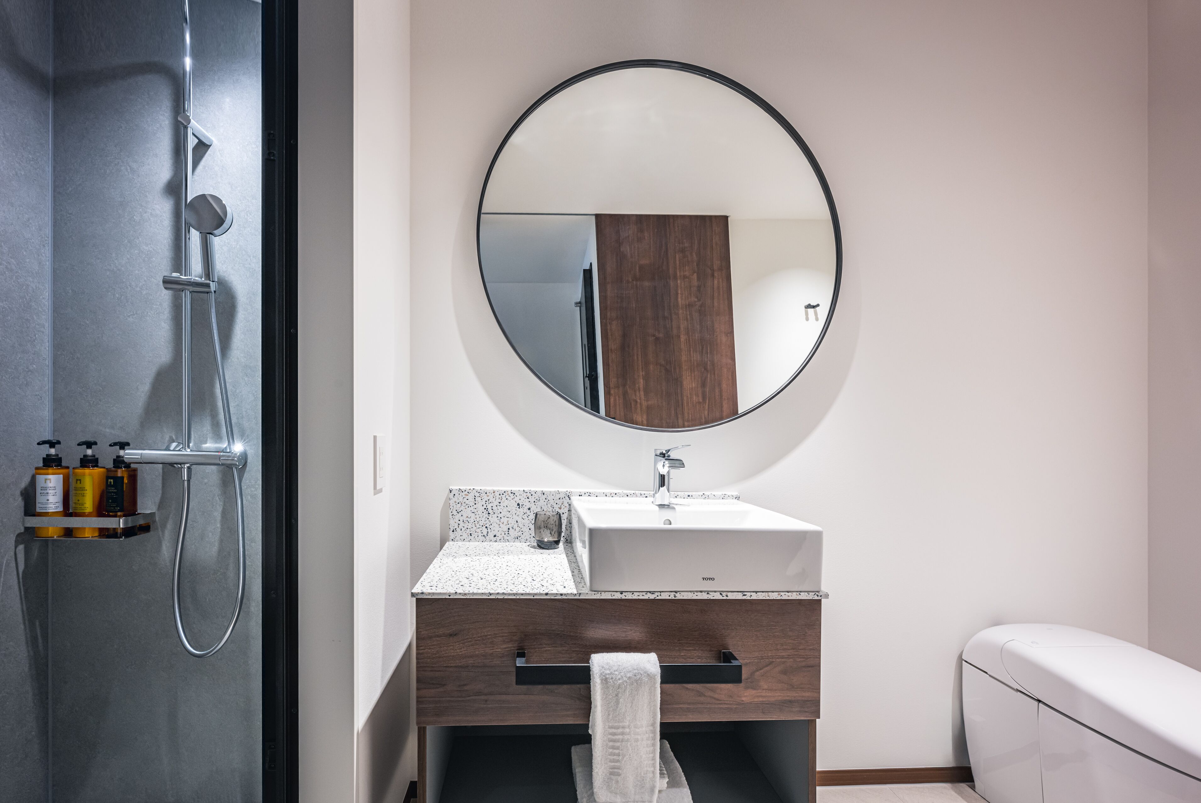 A modern bathroom with a freestanding shower, a round mirror, a white sink, and a wooden vanity.