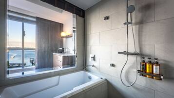A modern bathroom with a large bathtub, a shower, and a window offering a view of the sea.