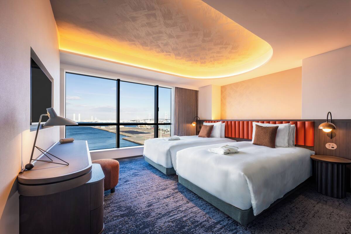 Une chambre d’hôtel moderne avec deux lits, une grande fenêtre donnant sur la ville, un bureau avec une lampe et une petite table d’appoint ronde.