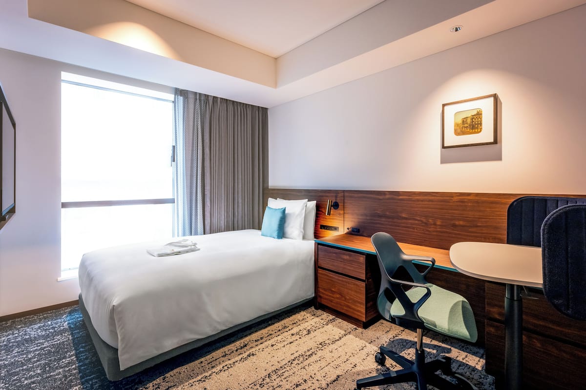 Une chambre d’hôtel avec un lit, un bureau, une chaise, une petite table, un tableau au mur et une fenêtre avec des rideaux.