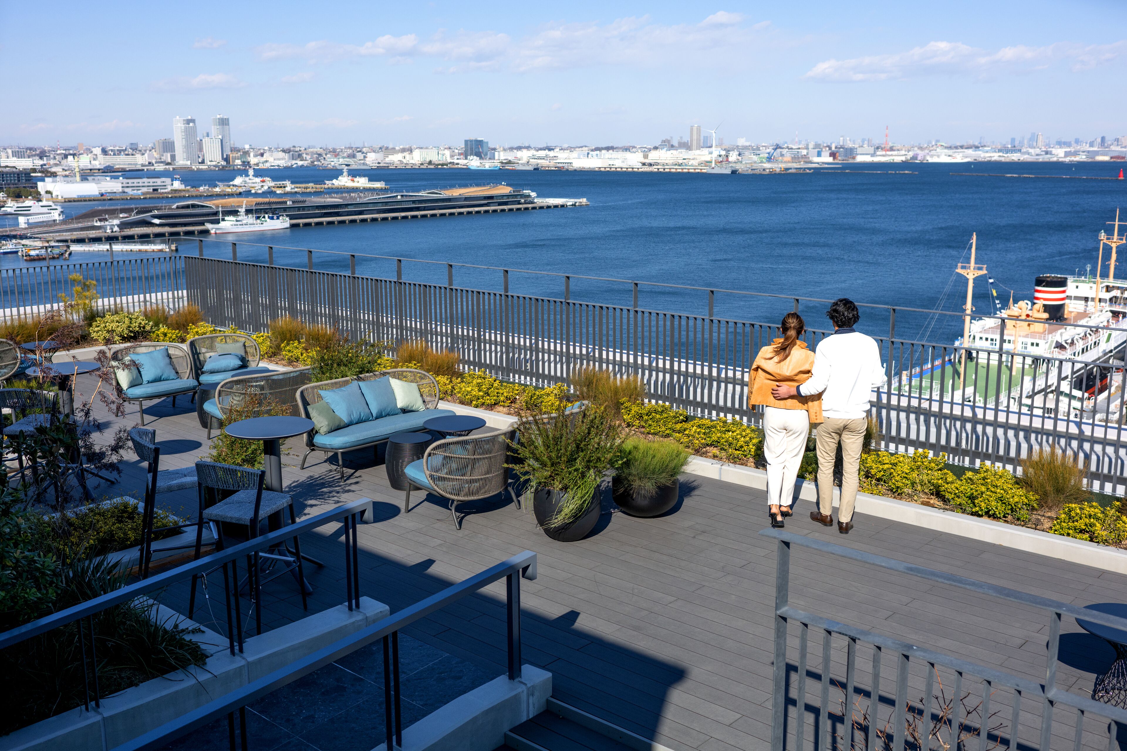 Una terraza en la azotea con mobiliario exterior, vistas al agua y un paisaje urbano de fondo.