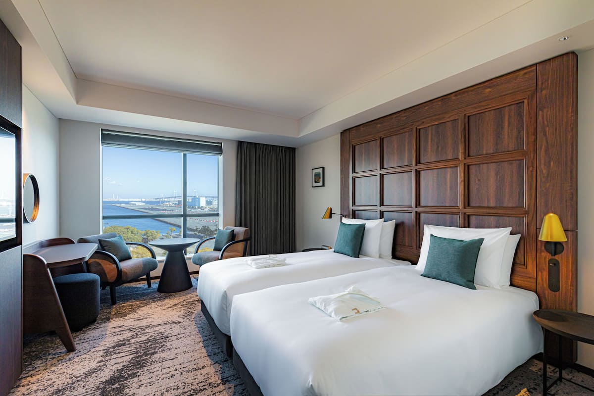 Une chambre d’hôtel avec deux lits, un bureau, une chaise, une petite table et une vue sur la ville.