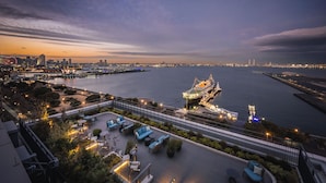 Ein Hotel am Wasserufer mit Außenbestuhlung, Blick auf die Skyline der Stadt und einem großen Schiff im Hafen.