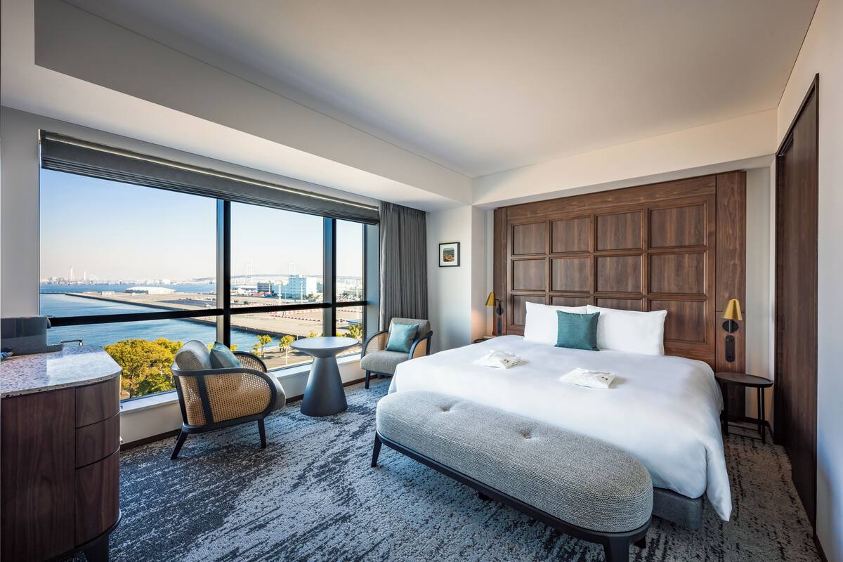 Une chambre d’hôtel avec un grand lit, un bureau, une chaise, une petite table et une vue sur la ville et l’eau.