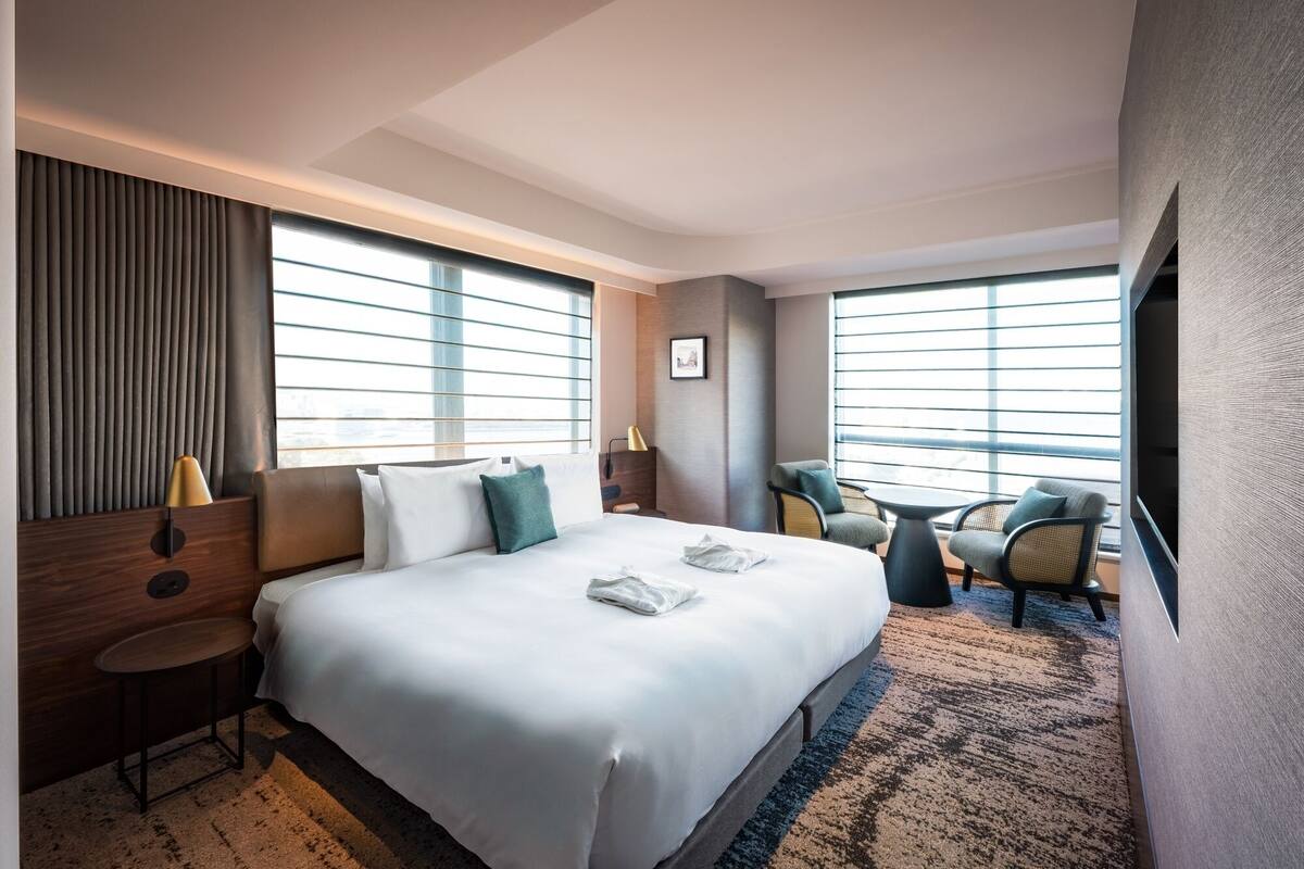 Une chambre d’hôtel avec un grand lit, une petite table, deux chaises, une télévision et de grandes fenêtres équipées de stores.