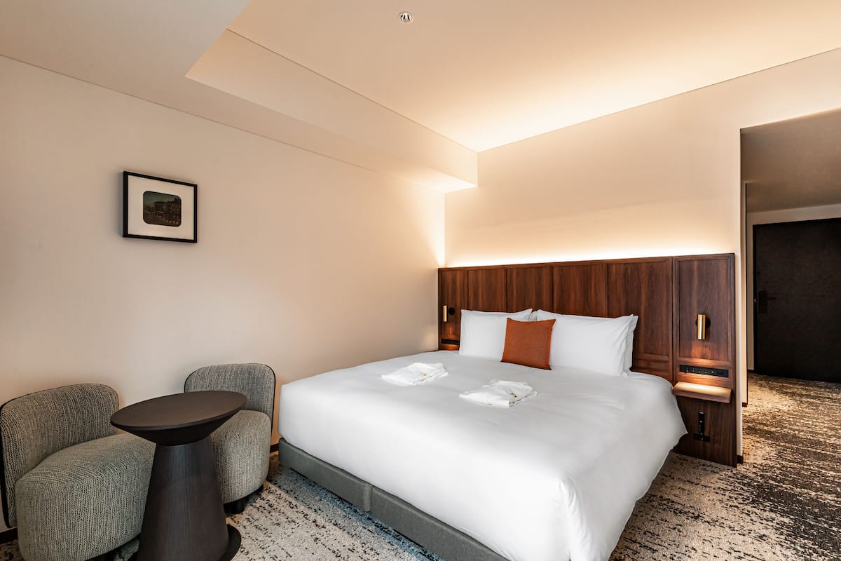 Une chambre d’hôtel avec un grand lit, une petite table ronde, deux fauteuils et un tableau encadré au mur.