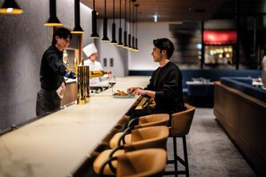 L’intérieur d’un restaurant moderne, avec un long comptoir, des tabourets de bar, des luminaires suspendus et un chef qui sert un client.