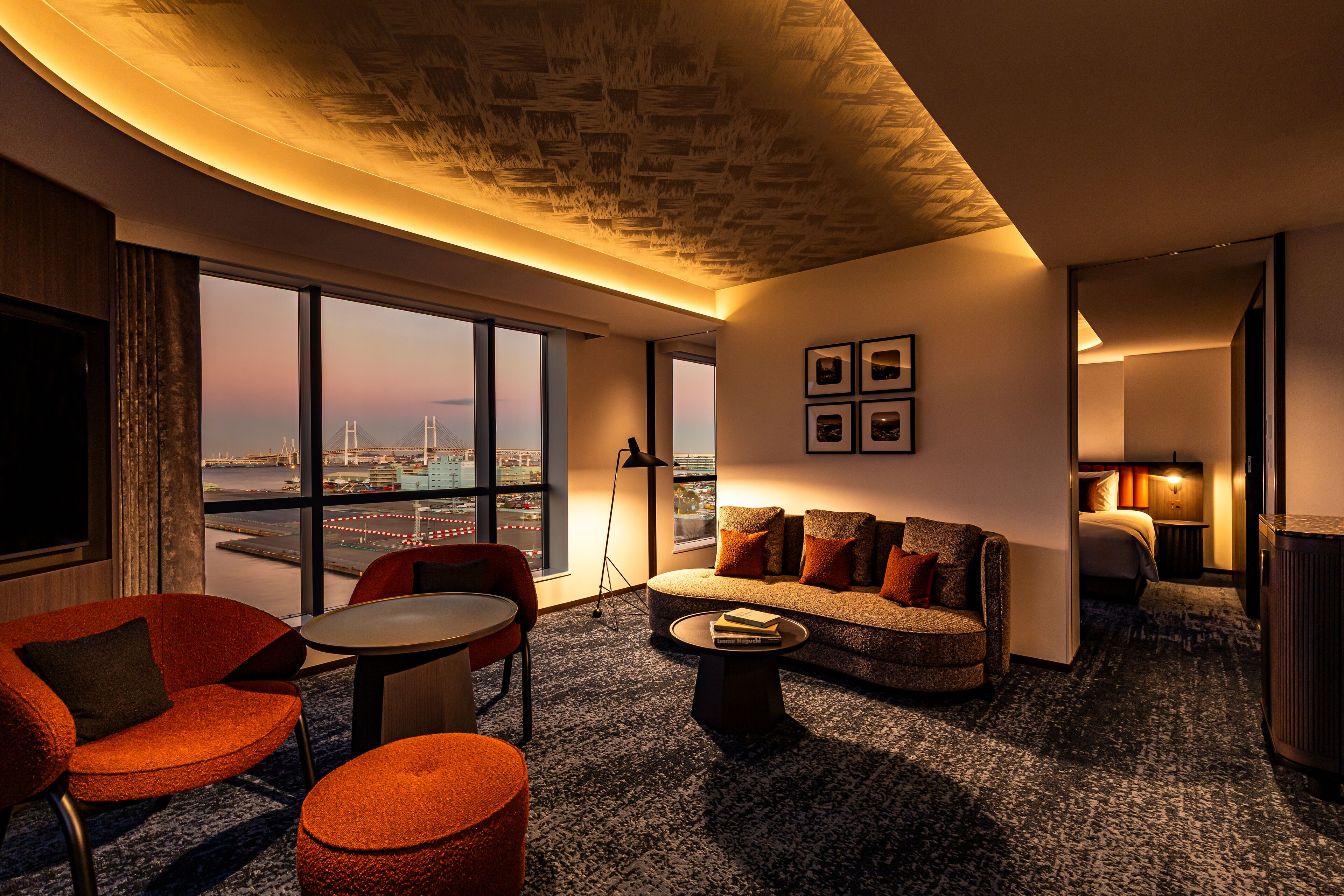 A modern hotel room with a curved ceiling, a large window offering a city view, a sofa, a chair, a small table, and a lamp.